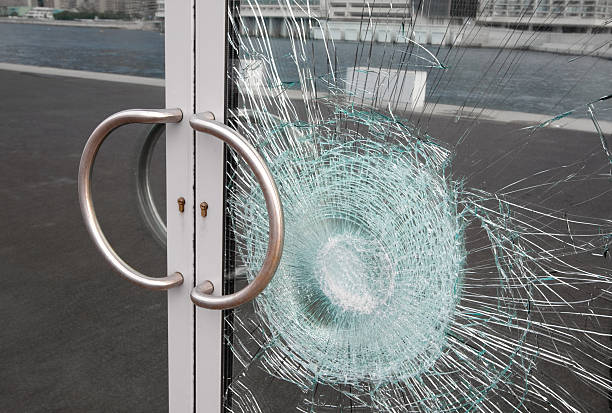 Broken glass door panel with smashed area. Shattered window on business entrance of an office building facade cracked and damaged by vandalism. The doubled glass pane is silvered on the back reflecting the cracks and sidewalk.